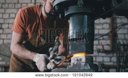 In the workshop of a blacksmith. A man with a beard puts a red-hot forged piece under an automatic hammer