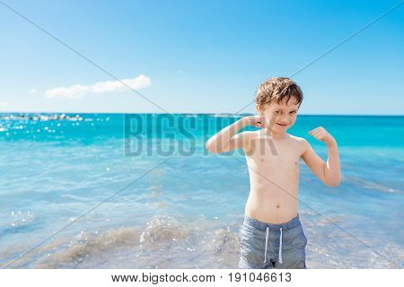 Confident Little Boy Showing His Muscles While Playing At The Beach