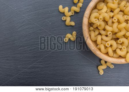Pasta makaroni in a wooden bowl on black background of slate or stone. Top view. Free space for your text