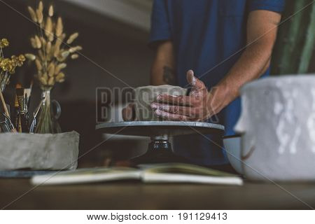 Close up of male hands working with clay on artisan working stand from dry wood surrounded by artistic workshop tools and implements creates poterry diy