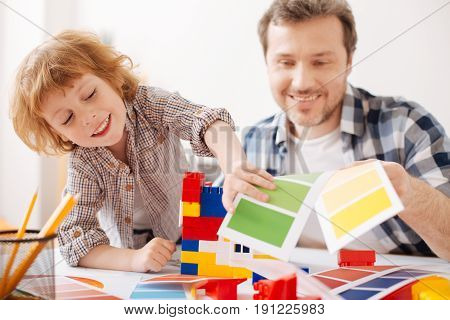 Stop working. Handsome boy leaning right elbow on the table while standing near his father and going to play with erector set