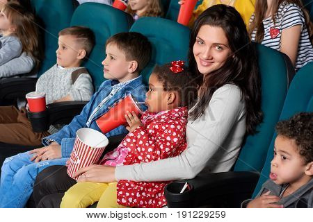 Cheerful young woman smiling to the camera happily with her little daughter sitting on her lap enjoying a movie at the cinema together family lifestyle leisure enjoyment happiness comedy cartoons.