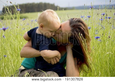 Happy family in the field of blew flowers. Beautiful young mother kisses her adorable baby boy. Concept of happiness.