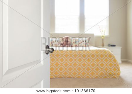 White door opened to sunny blurred defocused bedroom with big bed and windows in background in yellow and beige colors.