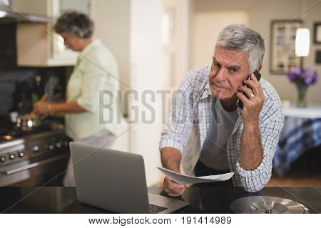 Portrait of senior man talking on phone while wife cooking in kitchen at home