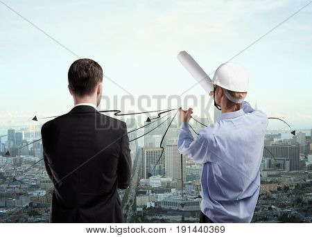 Back view of insdustrial worker explaining something to businessman on city background. Engineer concept