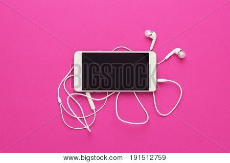 studio shot of smartphone and earbuds on purple background. top view of white smartphone and headphones. smartphone with white headset over bright pink. view from above of smartphone on bright colored back