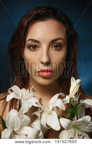 Closeup of gorgeous naked woman with wet hair posing in white lily flowers, looking straight.