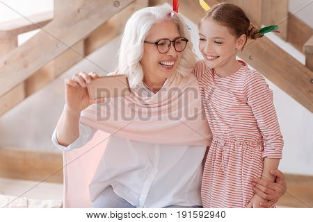 Lets take a selfie. Cheerful pleasant senior woman hugging her granddaughter and smiling while taking a selfie
