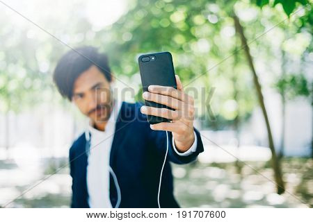 Handsome smiling businessman using smartphone for listining music while walking in city park.Young man making selfie portrait outdoor.Selective focus on hands.Horizontal, blurred background