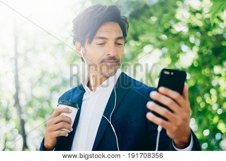 Closeup view of Handsome smiling businessman using smartphone for listining music while walking in city park.Young man making selfie portrait outdoor.Horizontal, blurred background