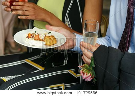 Guest holds a glass with champagne and a plate with desserts
