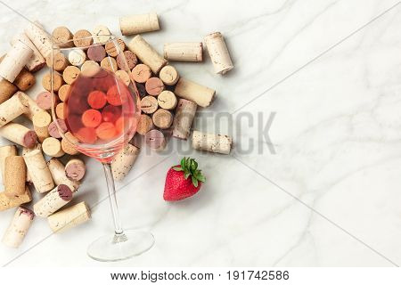 A high-key overhead photo of a glass of rose wine with corks and a fresh strawberry, on a white marble texture with a place for text