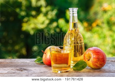 apple cider or juice in glass decanter with ripe fresh apples on wooden table with green natural background. Copy space. Summer drink