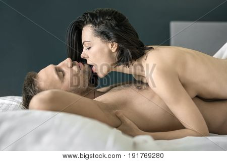 Hot nude couple with closed eyes lies on the white linens on the bed. Guy holds his hands under the head. Girl prepares to kiss him. Closeup. Indoors. Horizontal.