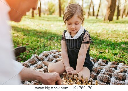 Anonymous senior man sitting on plaid with granddaughter playing chess in park.