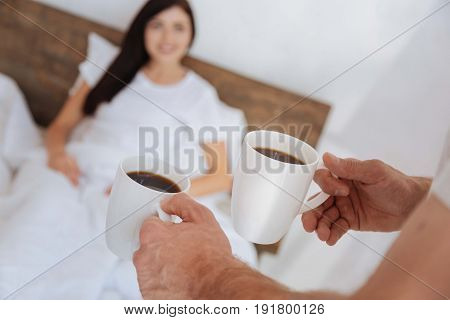 How did you sleep my love. Selective focus on male hands holding two cups full of coffee while bringing a breakfast for his girlfriend to their bed.