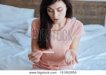 Cannot believe my eyes. Beautiful woman looking grievously at a positive pregnancy test while holding a handful of pills.
