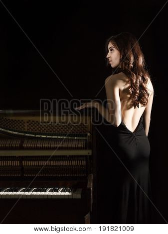 beautiful young woman in a black dress with an open back standing nere the piano on a dark background.