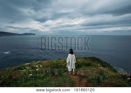 Young hipster nomad girl in casual clothing stands on edge of cliff contemplating and meditating finding peace and tranquility within herself