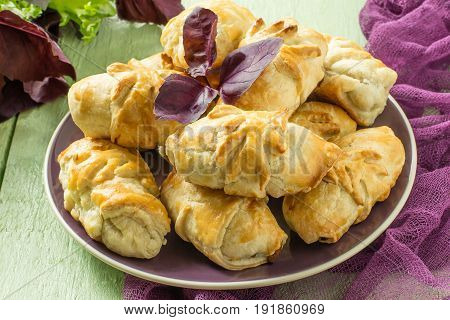 Delicious homemade rolls with eggplant cheese parsley and dill. Baking from puff pastry with vegetables. On green wooden table is a plate with pies on purple gauze napkin fresh lettuce and basil