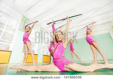Portrait of preteen rhythmic gymnasts performing front splits and doing exercise with clubs in school sports hall