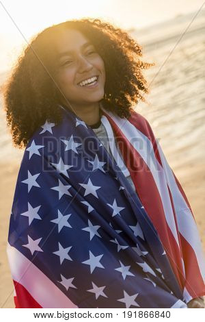 Beautiful happy mixed race African American girl teenager female young woman wrapped in the stars and stripes US flag on a beach smiling laughing with perfect teeth in golden sunset evening sunshine