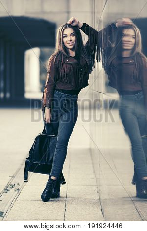 Happy young woman with handbag in city street. Stylish fashion model in leather fringe suede jacket and blue jeans