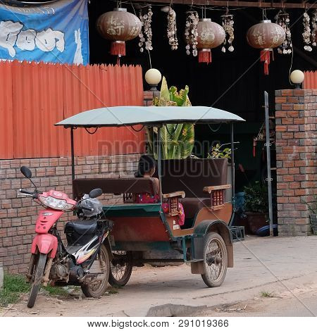 Cambodia, Siem Reap 12/08/2018 A Little Asian Girl Sits In A Moto Rickshaw Near A House With Red Lan