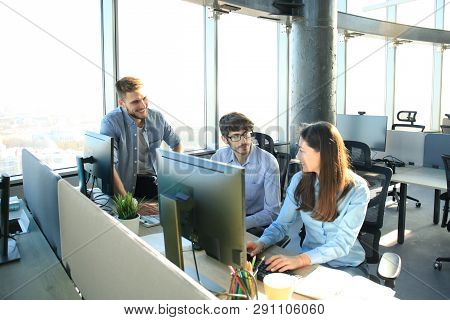 Group Of Young People In Casual Wear Sitting At The Office Desk And Discussing Something While Looki