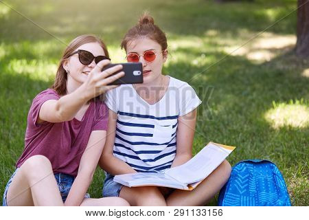 Outdoor Shot Of Two Young Beautiful Females Sitting On Grass In Lotus Position, Makes Selfie In Park