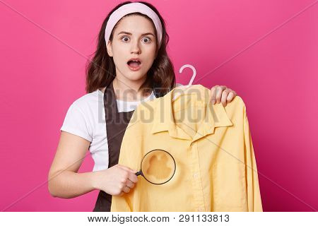 Image Of Shocked Female Wears White T Shirt, Brown Apron And Hair Band, Holds Yellow Shirt And Magni