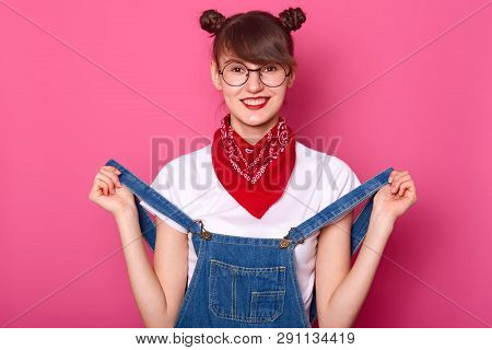 Portrait Of Smiling Female With Funny Bunches, Wears T Shirt, Denim Overalls And Bandana On Neck, Ha
