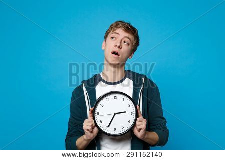 Portrait Of Preoccupied Young Man In Casual Clothes Holding Round Clock Isolated On Blue Wall Backgr