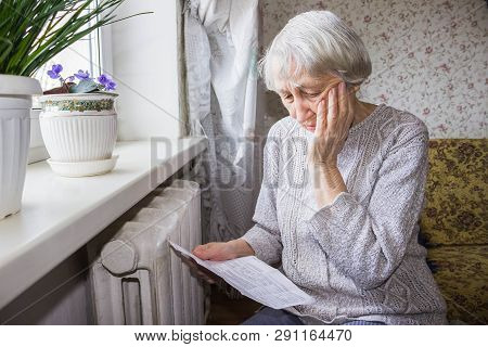 The Senior Woman Holding Gas Bill In Front Of Heating Radiator. Payment For Heating In Winter.