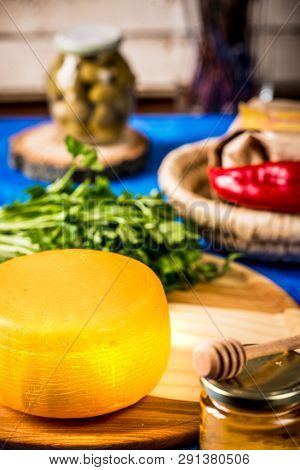Cheese Wheel On A Wooden Board Lies On The Table With Food And Fresh Herbs. Blurred Background.