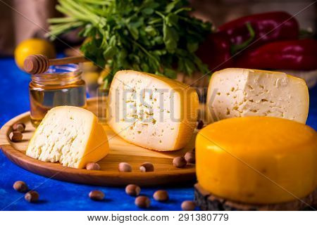 Cheese Wheel On A Wooden Board Lies On The Table With Food And Fresh Herbs. Blurred Background.