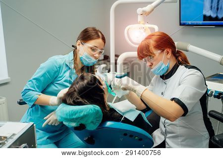 Stomatologist And Assistant Women In Masks And Protective Glasses Working With A Female Patient In D