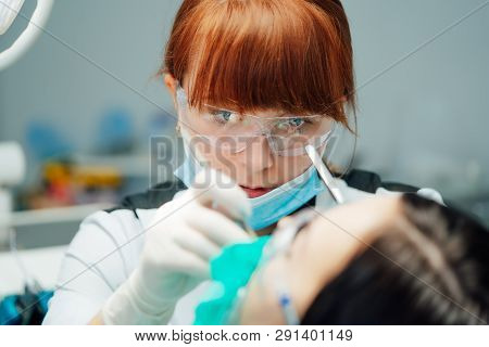 Red-haired Dentist In Safety Glasses And Mask Examining Patients Teeth In Clinic. Stomatologist Cari