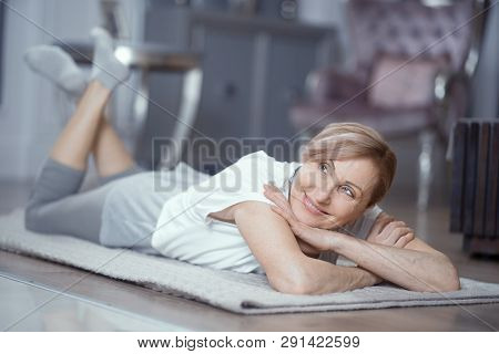 A Woman Over 50 Enjoys Relaxation After Yoga. She Lies On The Floor At Home. Woman Gently Smiles. Yo