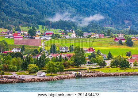 Norwegian Village Landscape With Turquoise Fjord Water, Mountains, Church And Colorful Houses. Loen,