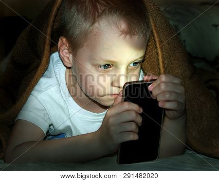Little Boy In Bed Under A Blanket Looking At The Smartphone At Night.