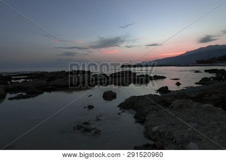 Pink And Blue Sunset On The Beach In Creta, Greece. Beach With Blacks Rocks And Waves At Dusk