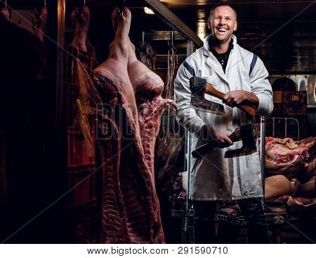 Cheerful Butcher In Workwear Posing With Two Axes In A Refrigerated Warehouse In The Midst Of Meat C