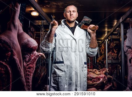 The Butcher In Workwear Posing With Two Axes In A Refrigerated Warehouse In The Midst Of Meat Carcas