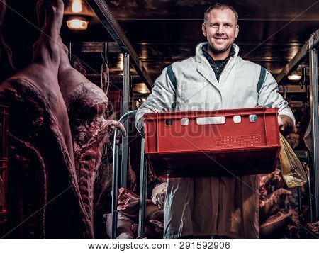 Cheerful Butcher In Workwear Holding A Box With Meat Pieces While Standing In The Midst Of Meat Carc