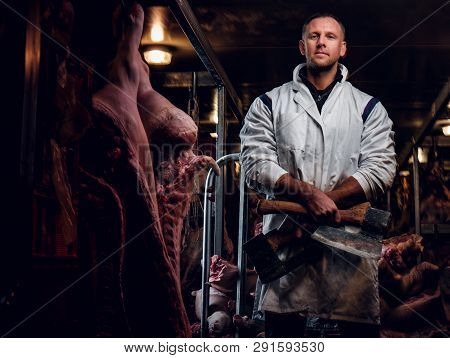 The Butcher In Workwear Posing With Two Axes In A Refrigerated Warehouse In The Midst Of Meat Carcas