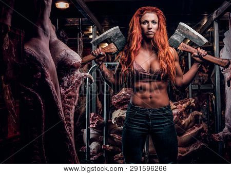 Redhead Fitness Woman In Bra And Jeans Posing With Two Axes In A Refrigerated Warehouse In The Midst