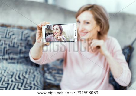 Elderly Woman Posing Taking Pictures Herself On The Phone. A Woman Is Sitting On The Sofa At Home. T