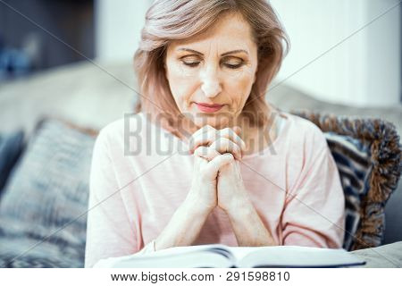 Middle Aged Woman Praying While Sitting On Sofa. Reading Pray From Bible. Religion Concept.